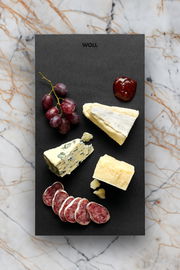 Frühstücksset mit Käse, Wurst, Trauben und Marmelade auf einer schwarzen Platte, von oben fotografiert.