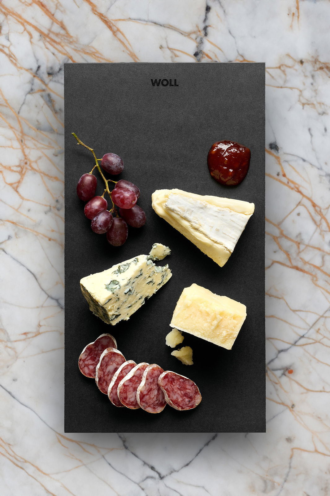 Frühstücksset mit Käse, Wurst, Trauben und Marmelade auf einer schwarzen Platte, von oben fotografiert.