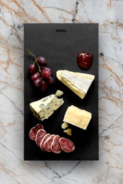 Frühstücksset mit Käse, Wurst, Trauben und Marmelade auf einer schwarzen Platte, von oben fotografiert.