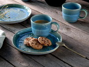 Schräg von oben fotografiertes Stillleben: blaue Keramik-Kaffeetasse auf passendem Teller mit drei Schokokeksen, daneben Löffel und weitere Tasse auf rustikalem Holztisch.