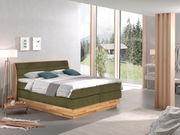 Boxspringbett in einem modernen Schlafzimmer mit großen Fenstern, die einen Blick auf die Natur bieten. Das Bett hat ein olivgrünes Kopfteil und eine Holzbasis. Perspektive von der Seite.
