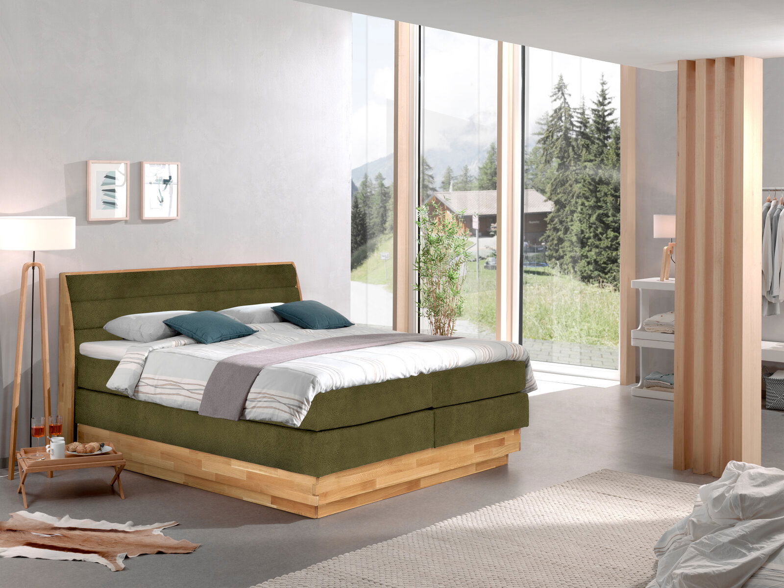 Z2 Boxspringbett MARISSA Boxspringbett in einem modernen Schlafzimmer mit großen Fenstern, die einen Blick auf die Natur bieten. Das Bett hat ein olivgrünes Kopfteil und eine Holzbasis. Perspektive von der Seite.