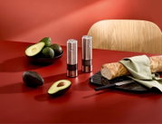 Zwei elektrische Salz- und Pfeffermühlen aus gebürstetem Edelstahl auf roter Küchenfläche, daneben Avocados und Baguette auf Schneidebrett; Perspektive: leicht schräg von vorn (3/4-Ansicht)
