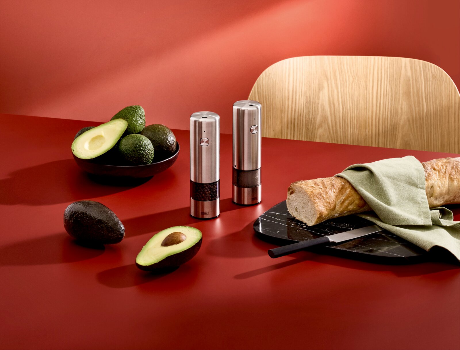 Zwei elektrische Salz- und Pfeffermühlen aus gebürstetem Edelstahl auf roter Küchenfläche, daneben Avocados und Baguette auf Schneidebrett; Perspektive: leicht schräg von vorn (3/4-Ansicht)