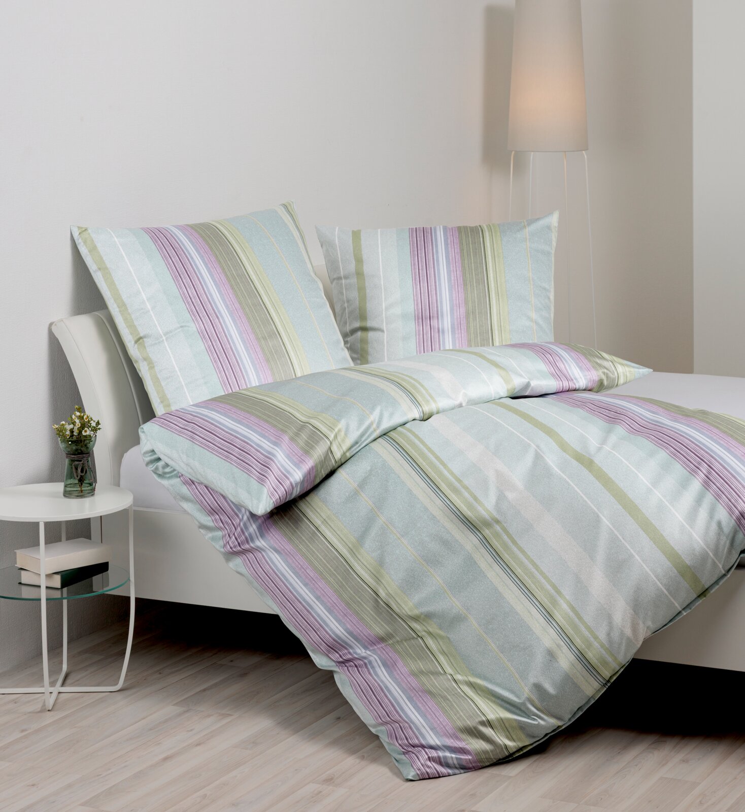 Janine Beddengoed Gestreepte beddengoed in pasteltinten (mint, groen, paars en wit) op een bed, schuin van voren gefotografeerd; een dekbedovertrek en twee kussenslopen in een slaapkameromgeving.