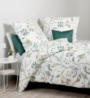 Bettwäsche-Set in Weiß mit botanischem Blätter- und Zweig-Muster in Grün und Akzenten in Orange, dekoriert mit dunkelgrünen Kissen, fotografiert in leichter Schrägansicht von vorne auf einem Bett im hellen Schlafzimmer.