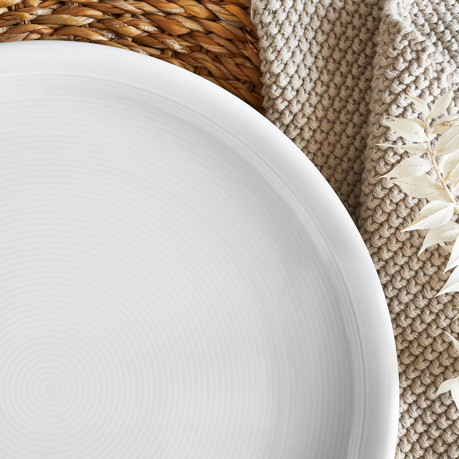 Nahaufnahme eines weißen Tellers mit feinen konzentrischen Kreisen, auf einem geflochtenen Untersetzer und einer beigen Textilunterlage, dekoriert mit getrockneten weißen Blättern.