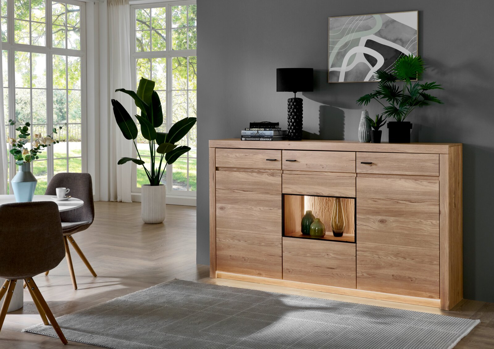 Cantus Sideboard WIEN Massives Sideboard aus hellem Holz mit Schubladen, Türen und beleuchtetem offenen Fach, dekoriert mit Lampe, Büchern und Pflanzen, fotografiert in leicht schräger Frontalansicht in einem modernen Wohnraum.