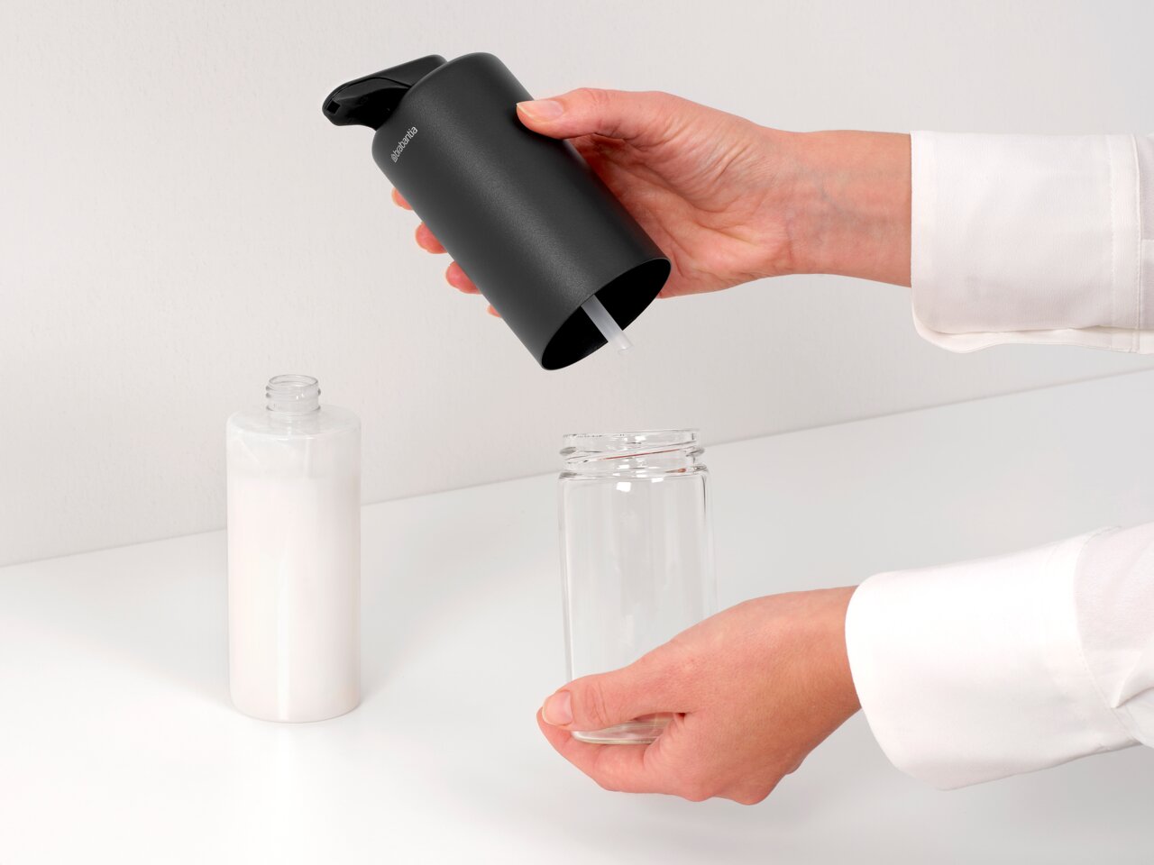 Nahaufnahme eines schwarzen Seifenspenders, der von einer Hand gehalten wird, während Seife in ein Glasgefäß gefüllt wird. Der Spender hat ein modernes Design und wird von der Seite gezeigt.