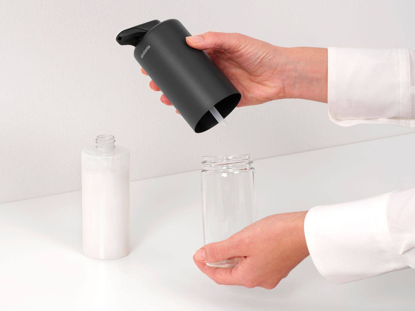 Nahaufnahme eines schwarzen Seifenspenders, der von einer Hand gehalten wird, während Seife in ein Glasgefäß gefüllt wird. Der Spender hat ein modernes Design und wird von der Seite gezeigt.