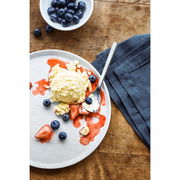 Vogelperspektive auf einen Teller mit einer Kugel Vanilleeis, umgeben von Erdbeersauce und frischen Blaubeeren, daneben eine Eisschaufel.