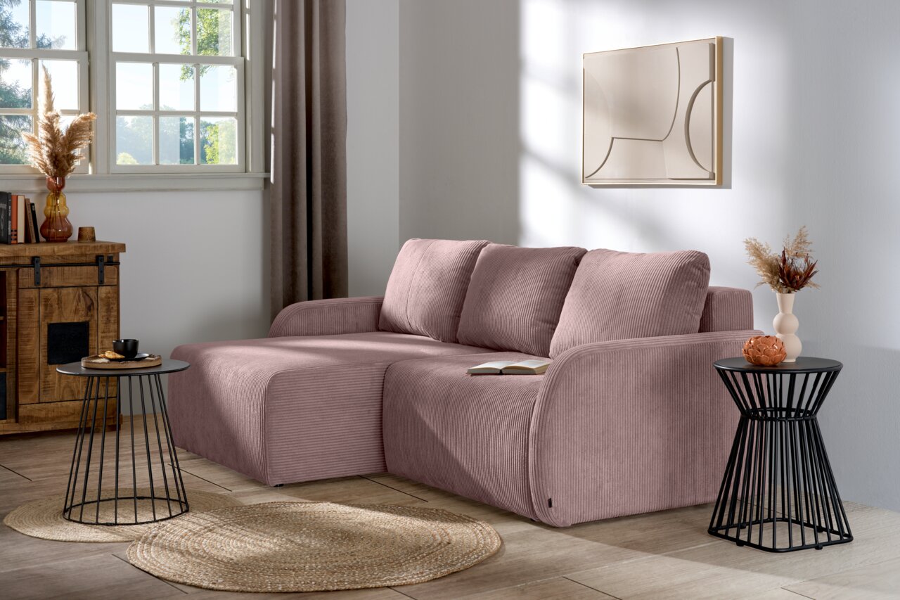 L-förmiges Sofa in einem modernen Wohnzimmer, aus einer seitlichen Perspektive fotografiert. Das Sofa hat eine rosa Farbe und ist mit Kissen ausgestattet. Es steht auf einem Holzboden neben einem runden Beistelltisch und einer Kommode.