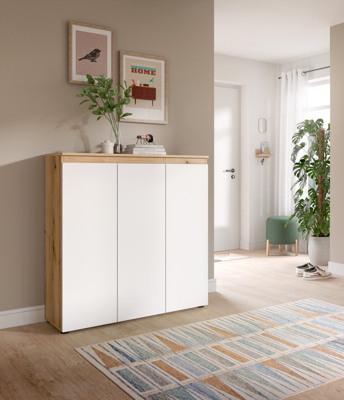 Modernes Sideboard/Schuhschrank in Weiß mit Holzrahmen und Holzplatte, drei Türen, im Flur dekoriert mit Pflanzen und Bildern Frontansicht aus leicht schräger Perspektive von links.