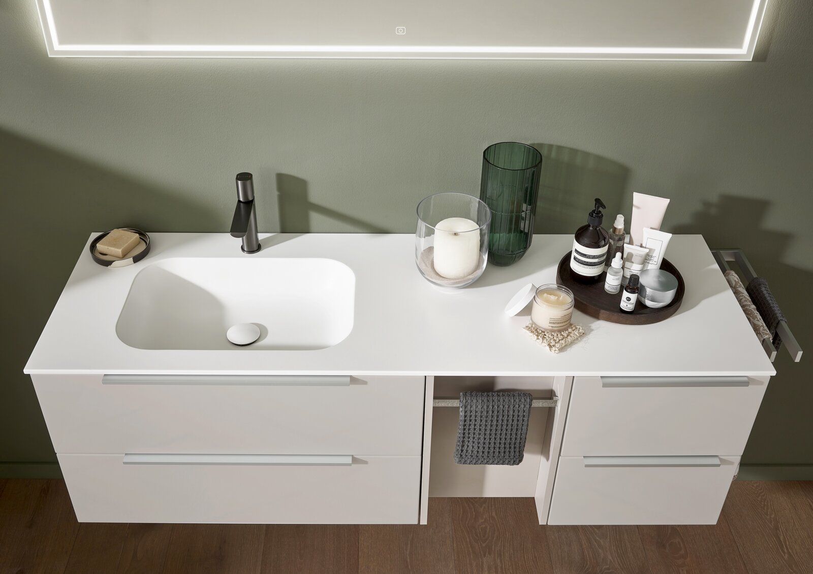 Modernes weißes Badezimmer-Waschtischmöbel mit integrierter Waschschale und schwarzer Armatur, dekoriert mit Kerze, Vase und Pflegeprodukten, Ansicht von vorne leicht von oben