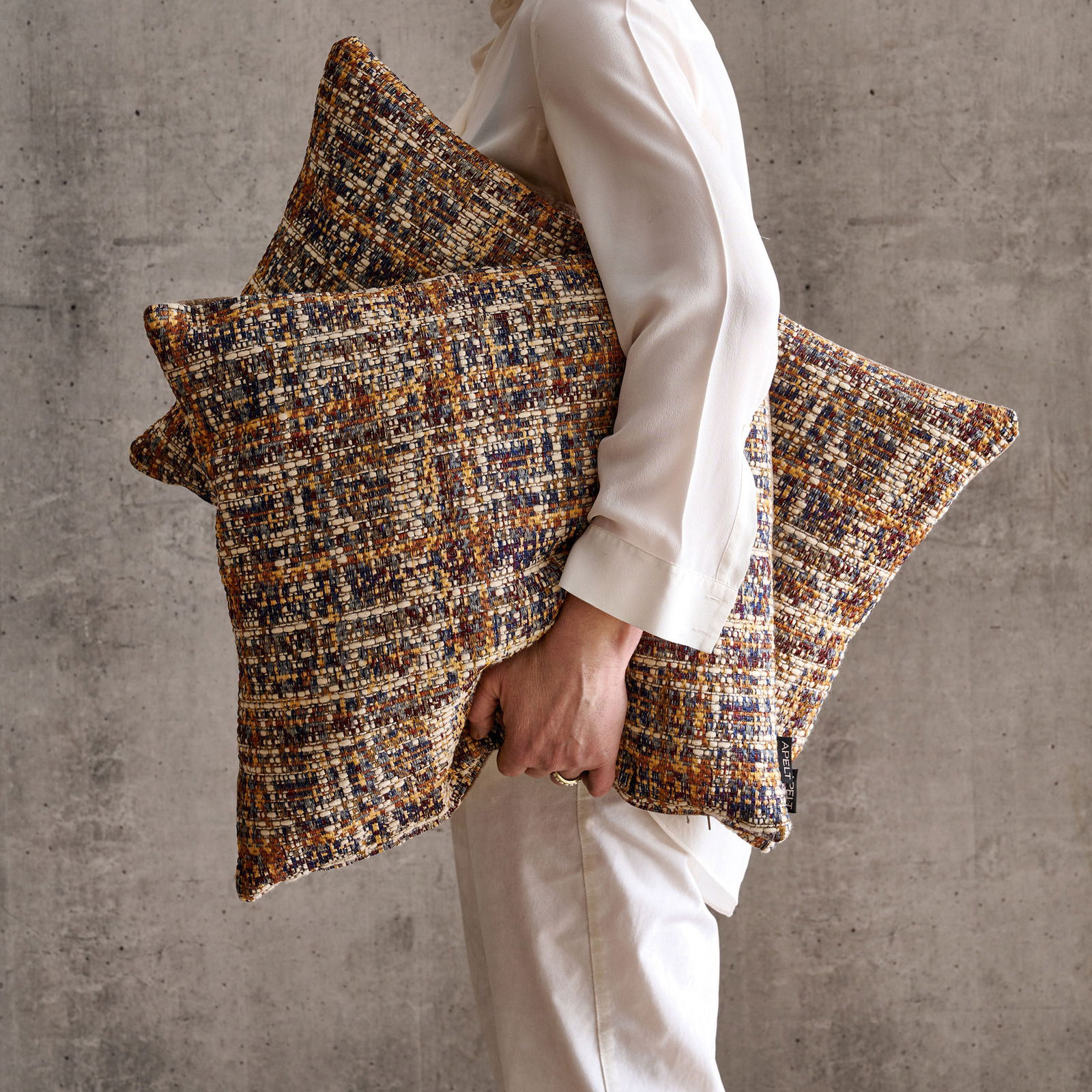 Person in weißem Outfit hält zwei Wendekissenhüllen mit buntem, texturiertem Muster vor einer grauen Wand, seitliche Perspektive.