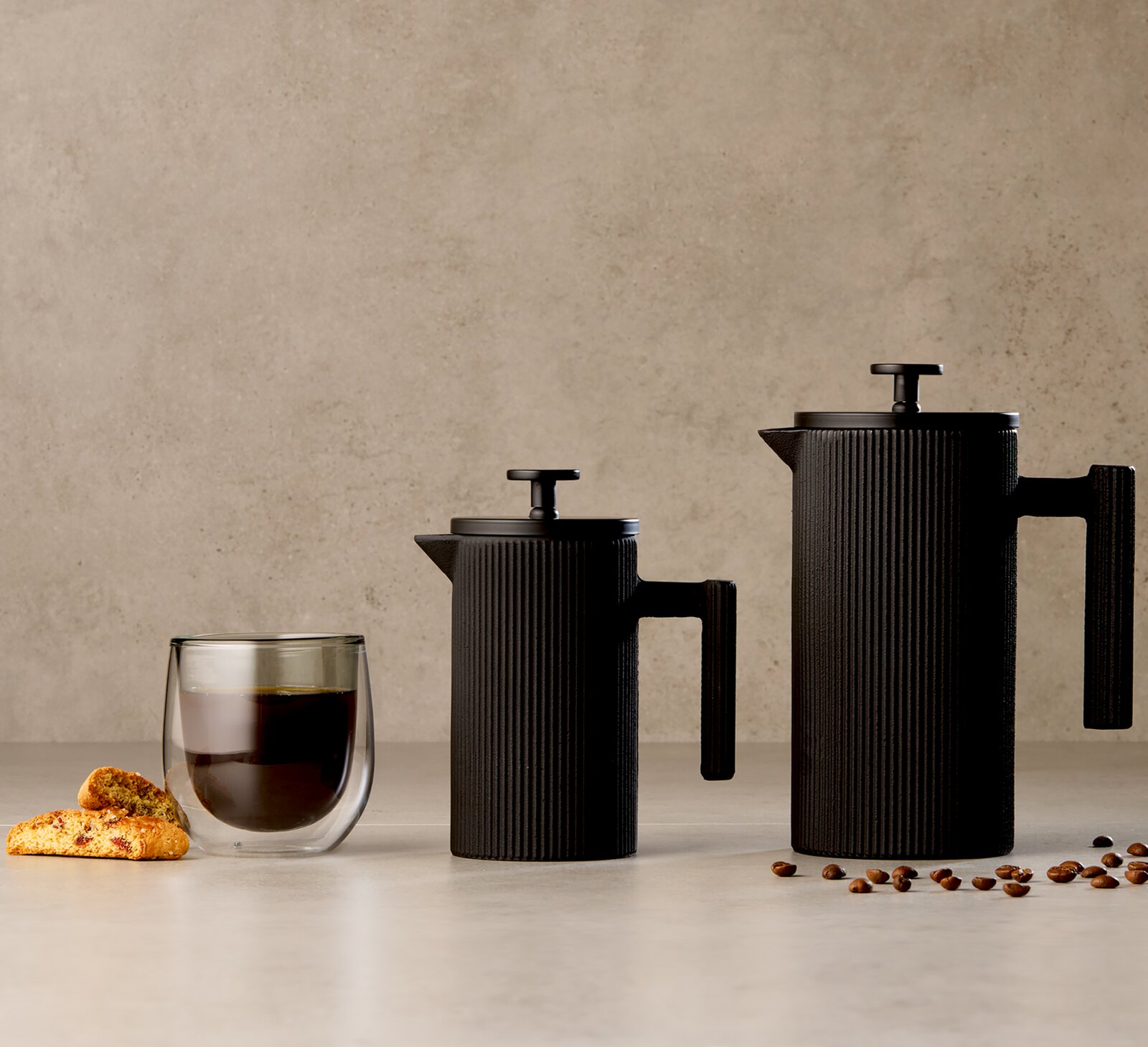 Zwei schwarze, geriffelte French-Press-Kaffeebereiter in unterschiedlichen Größen neben einem doppelwandigen Kaffeeglas, Gebäck und Kaffeebohnen, frontal aus leichter Seitenperspektive präsentiert
