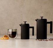 Zwei schwarze, geriffelte French-Press-Kaffeebereiter in unterschiedlichen Größen neben einem doppelwandigen Kaffeeglas, Gebäck und Kaffeebohnen, frontal aus leichter Seitenperspektive präsentiert