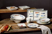 Maritimes Geschirrset aus weißer Keramik mit bunten Fischmotiven und blauen Randlinien, bestehend aus Tellern, Schalen, Tasse und eckigen Platten, in schräger Frontansicht auf einem rustikalen Holztisch arrangiert.
