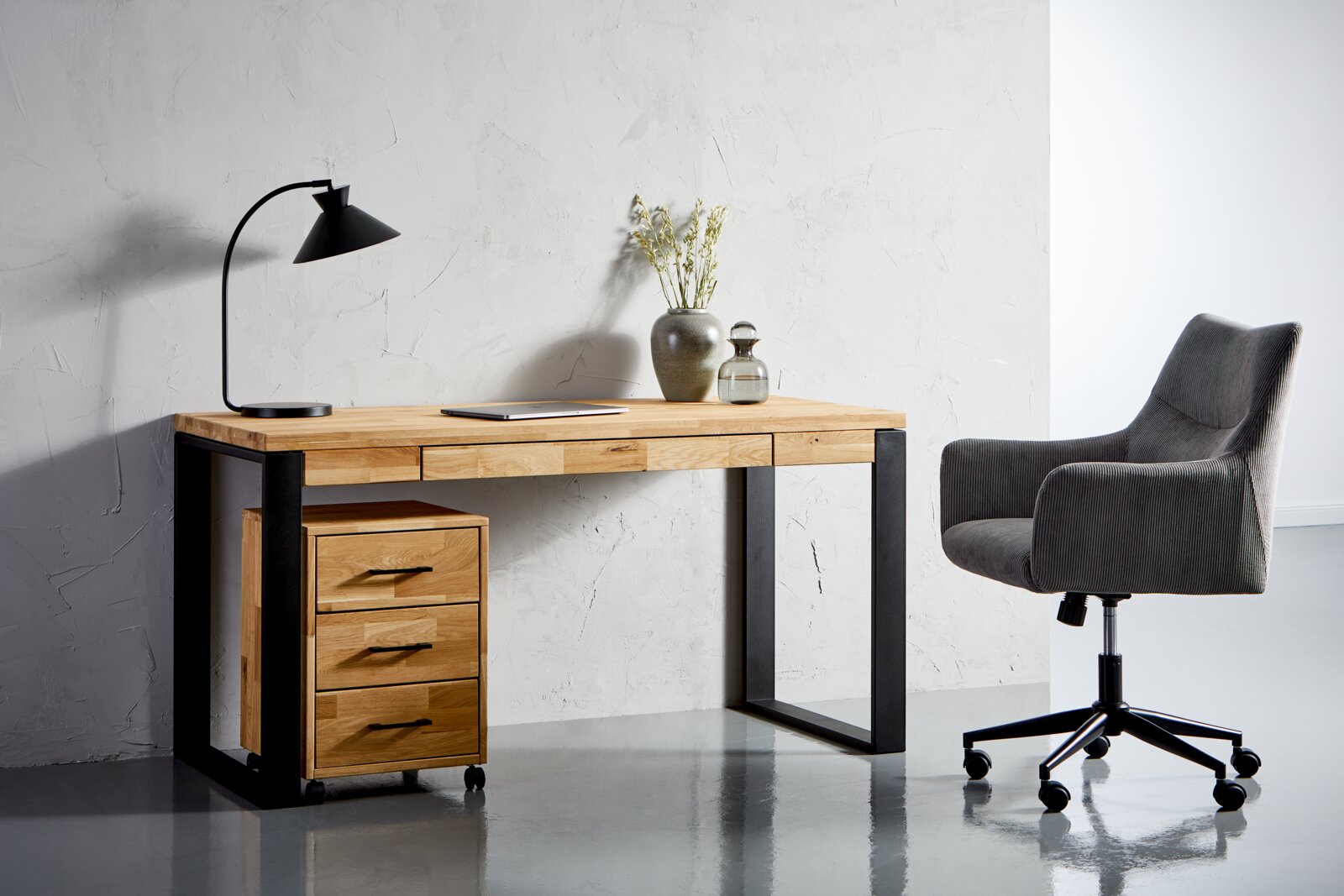 Moderner Schreibtisch aus Holz mit schwarzem Metallgestell, ausgestattet mit einer Schubladeneinheit auf Rollen, einer Tischlampe und Dekorationen, seitliche Perspektive.