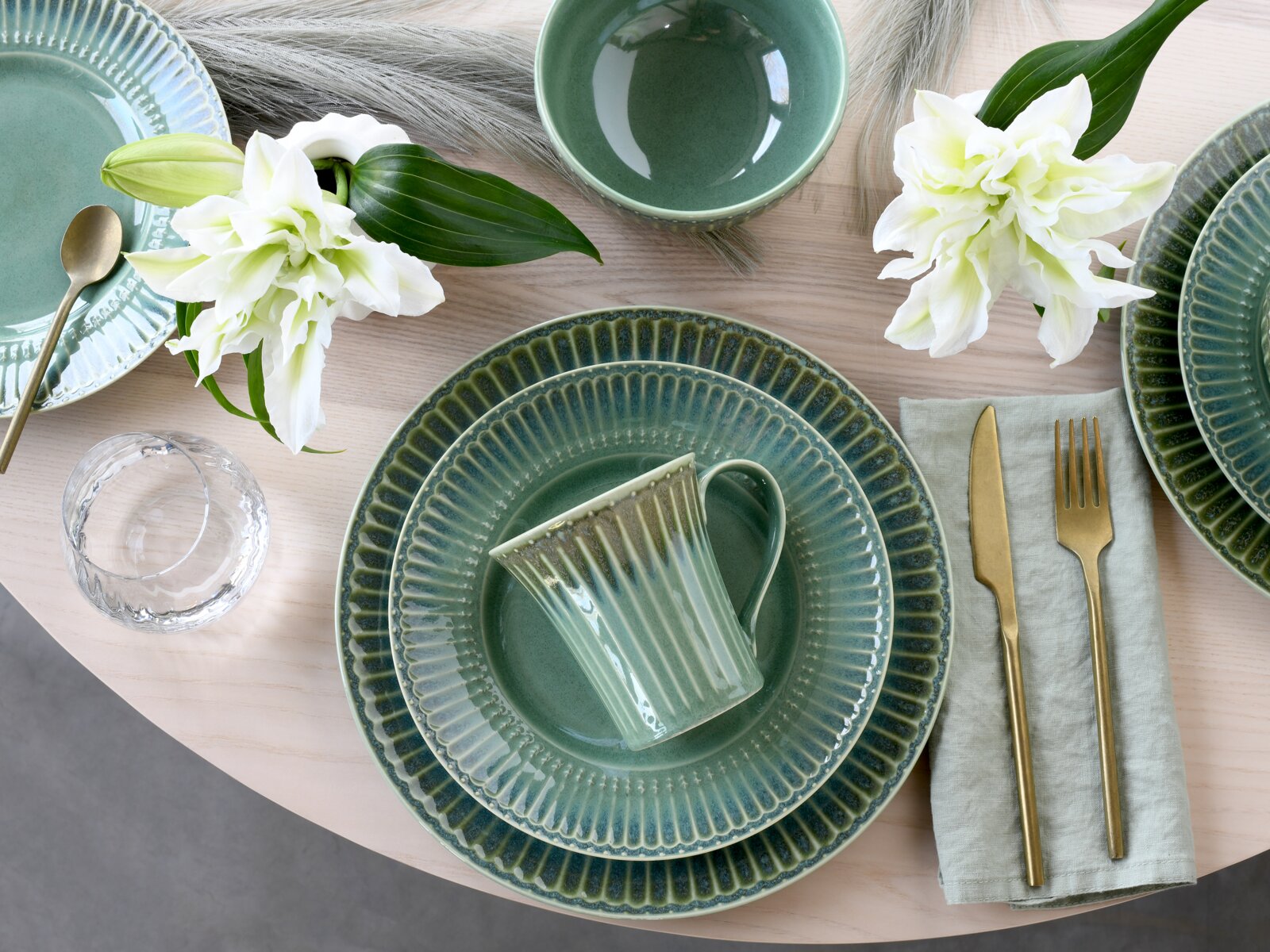 Draufsicht auf ein stilvoll gedecktes Tischarrangement mit grün glasiertem Keramikgeschirr (Teller, Schale und geriffelter Tasse), goldfarbenem Besteck auf hellgrüner Serviette, Wasserglas und weißen Lilien auf hellem Holztisch.