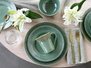 Draufsicht auf ein stilvoll gedecktes Tischarrangement mit grün glasiertem Keramikgeschirr (Teller, Schale und geriffelter Tasse), goldfarbenem Besteck auf hellgrüner Serviette, Wasserglas und weißen Lilien auf hellem Holztisch.