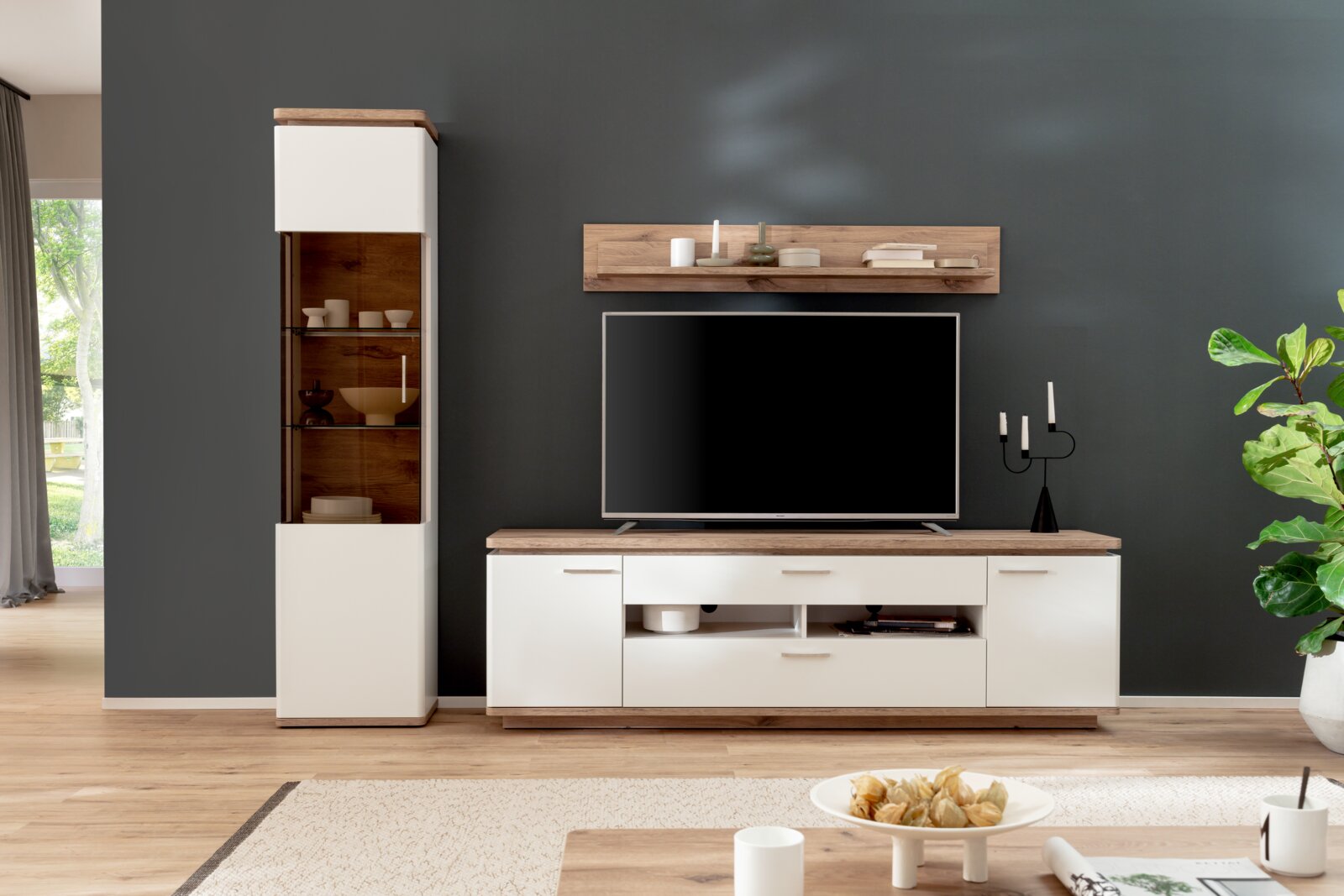 Wohnwand im modernen Design mit weißer TV-Kommode mit Holzoptik-Deckplatte, passendem Hängeregal und hoher Vitrine mit Glasfront, frontale Perspektive im Wohnzimmer-Setting