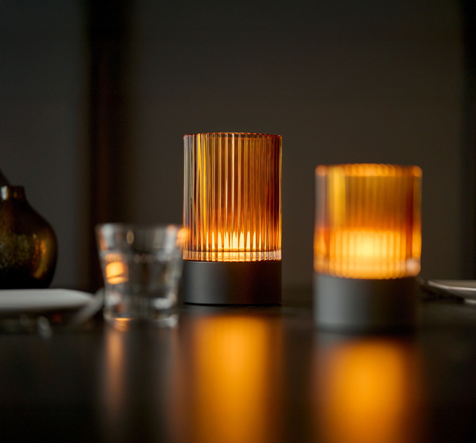 Zwei Akku-Windlichter auf einem Tisch, die warmes, stimmungsvolles Licht ausstrahlen. Perspektive: seitlich, leicht von unten.