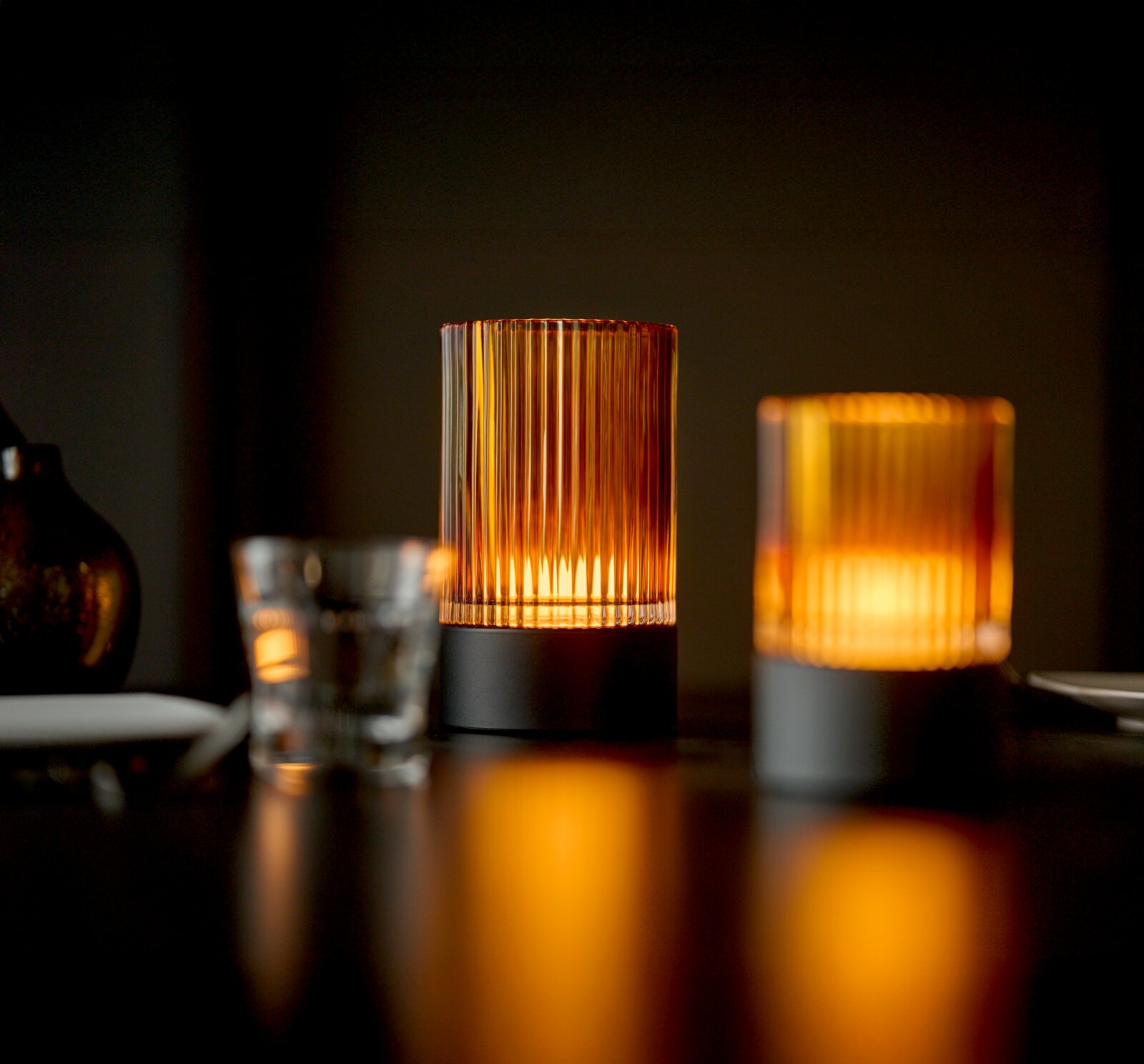 Zwei Akku-Windlichter auf einem Tisch, die warmes, stimmungsvolles Licht ausstrahlen. Perspektive: seitlich, leicht von unten.