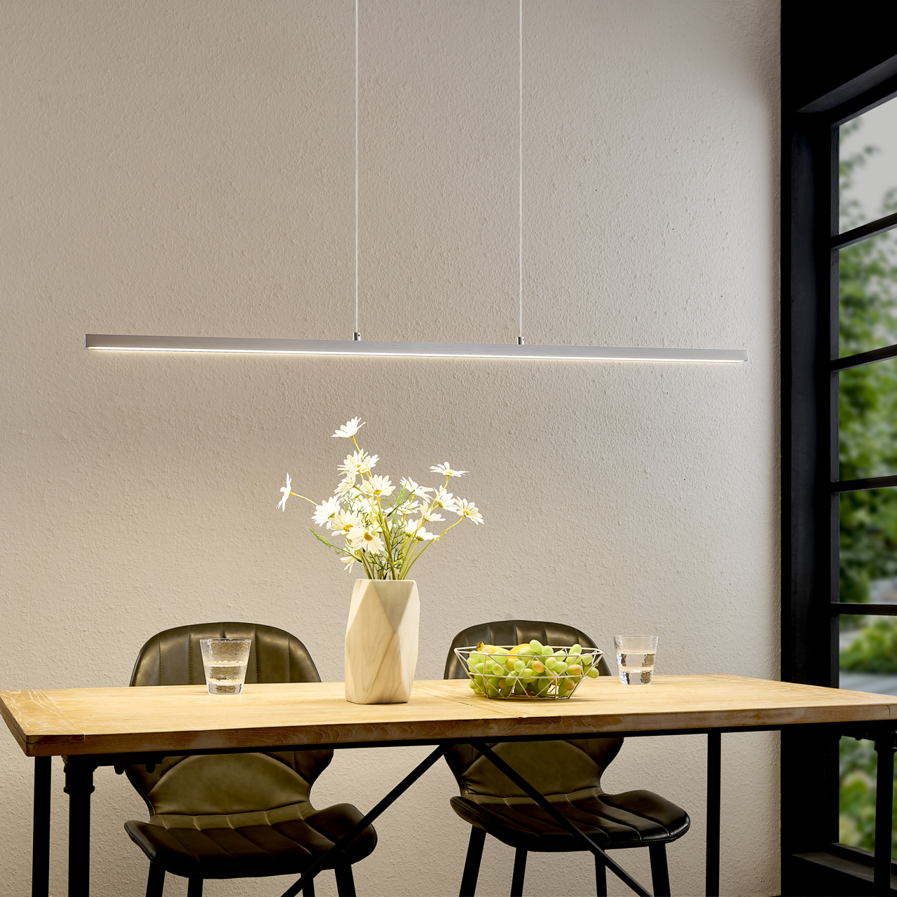 Moderne, schlanke LED-Pendelleuchte in Silber über einem Holztisch mit zwei Stühlen, Vase mit weißen Blumen und Obstschale; frontale Perspektive auf die Leuchte im Essbereich