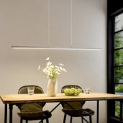Moderne, slanke LED-hanglamp in zilver boven een houten tafel met twee stoelen, een vaas met witte bloemen en een fruitschaal; vooraanzicht van de lamp in de eethoek