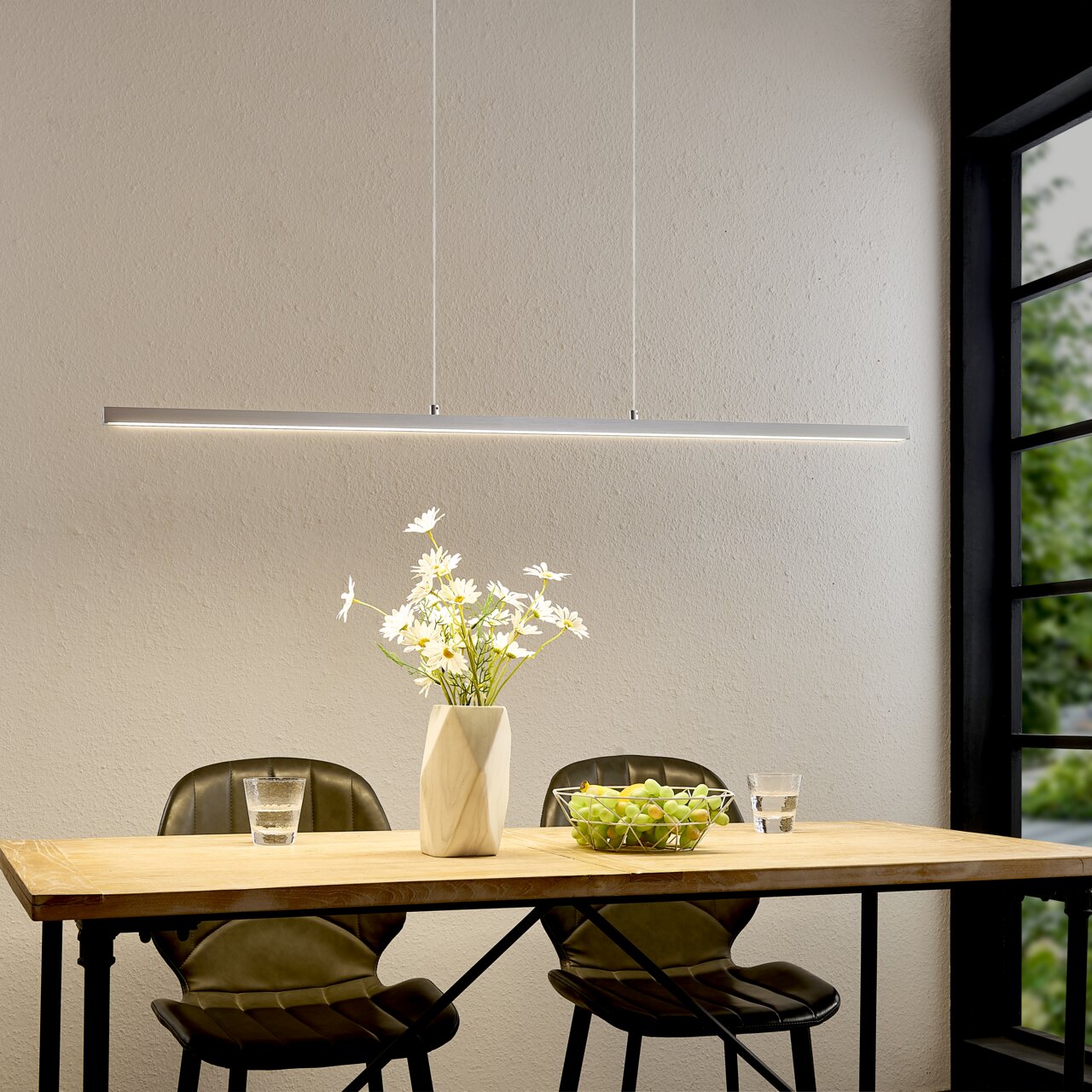 Moderne, slanke LED-hanglamp in zilver boven een houten tafel met twee stoelen, een vaas met witte bloemen en een fruitschaal; vooraanzicht van de lamp in de eethoek