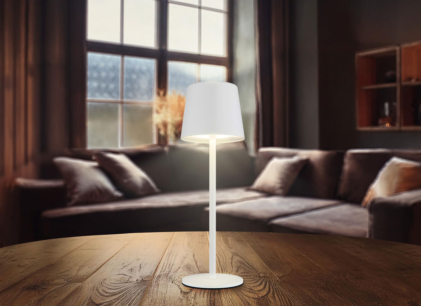 Moderne Tischleuchte mit weißem Schirm auf einem Holztisch, im Hintergrund ein gemütliches Wohnzimmer mit Sofa und großem Fenster, seitliche Perspektive.