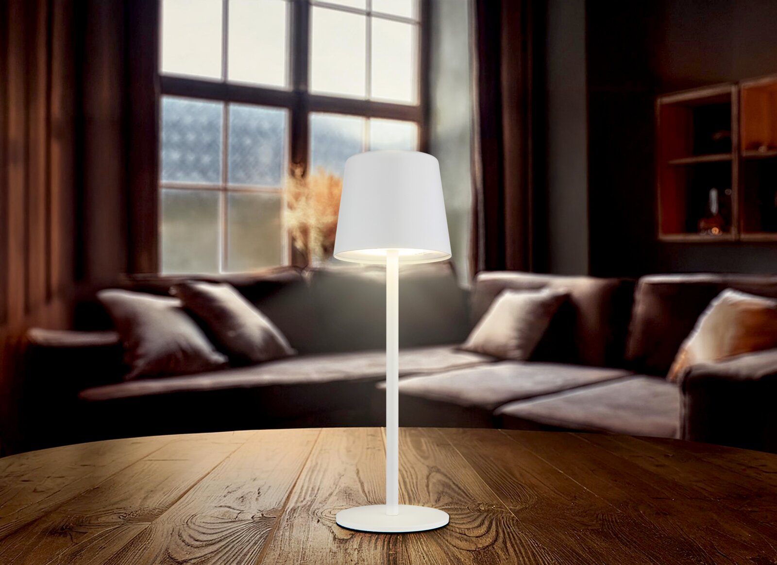 Moderne Tischleuchte mit weißem Schirm auf einem Holztisch, im Hintergrund ein gemütliches Wohnzimmer mit Sofa und großem Fenster, seitliche Perspektive.