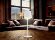 Moderne Tischleuchte mit weißem Schirm auf einem Holztisch, im Hintergrund ein gemütliches Wohnzimmer mit Sofa und großem Fenster, seitliche Perspektive.