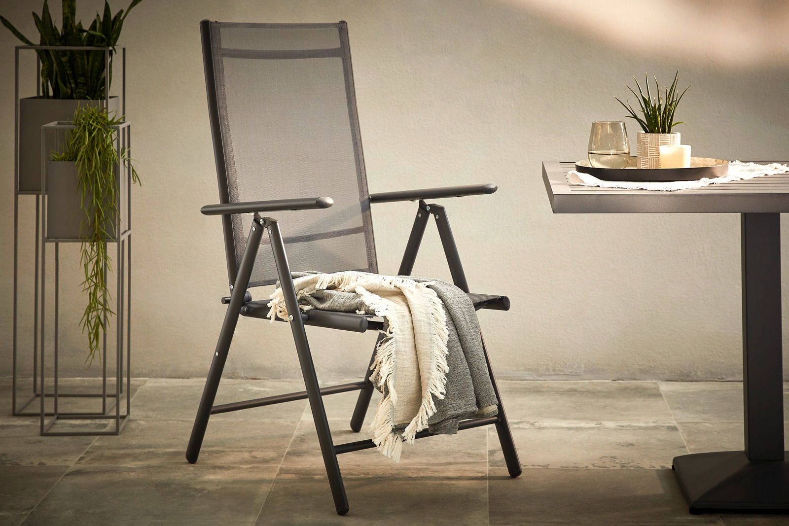 Moderner Gartenstuhl aus Metall mit Armlehnen, seitliche Perspektive, neben einem Tisch und Pflanzen.