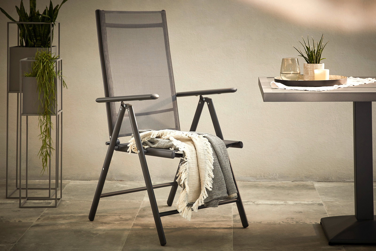 Moderner Gartenstuhl aus Metall mit Armlehnen, seitliche Perspektive, neben einem Tisch und Pflanzen.