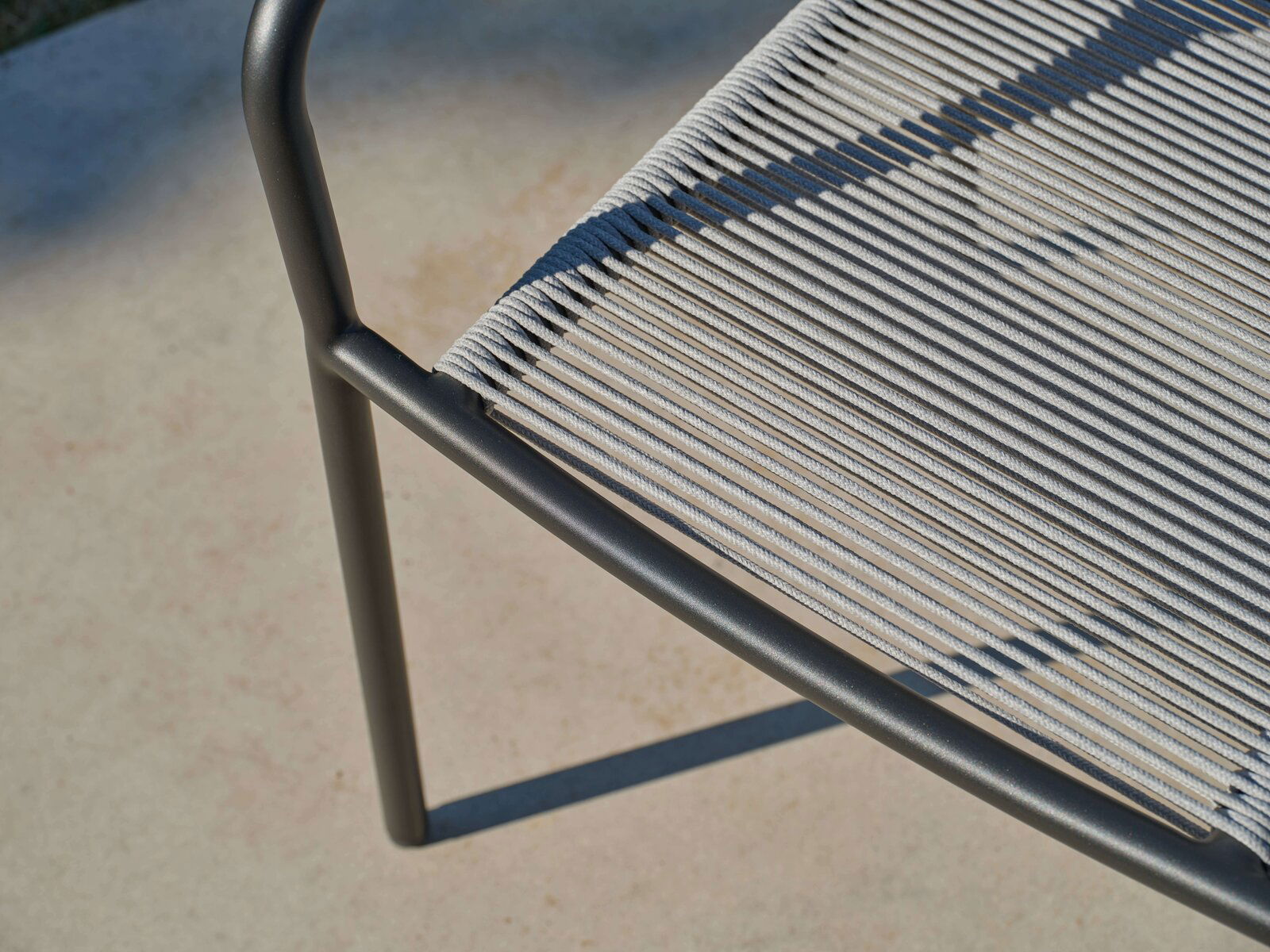 Nahaufnahme eines Stapelsessels mit schwarzem Metallrahmen und grauer Seilbespannung, von oben betrachtet.