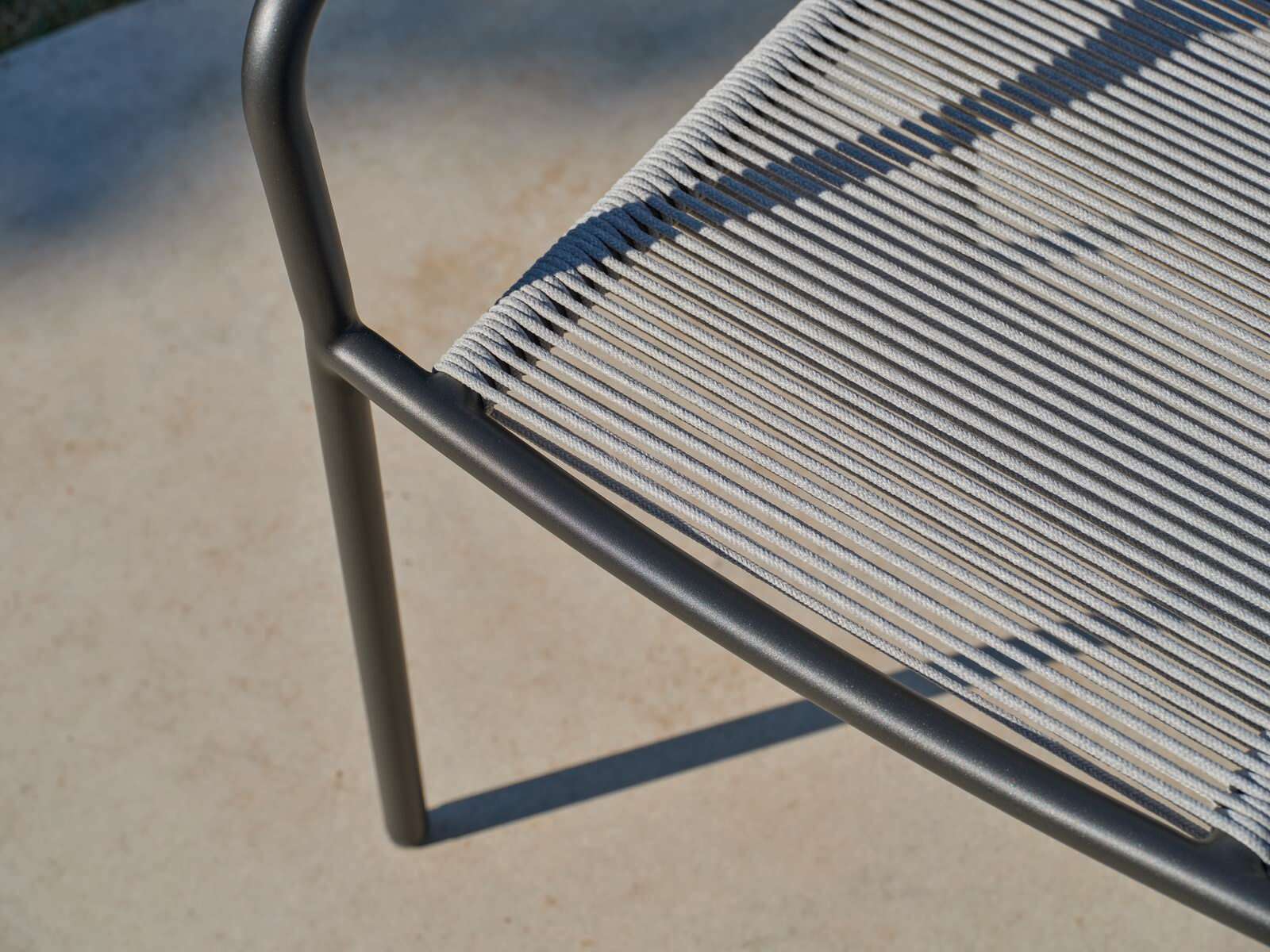 Nahaufnahme eines Stapelsessels mit schwarzem Metallrahmen und grauer Seilbespannung, von oben betrachtet.