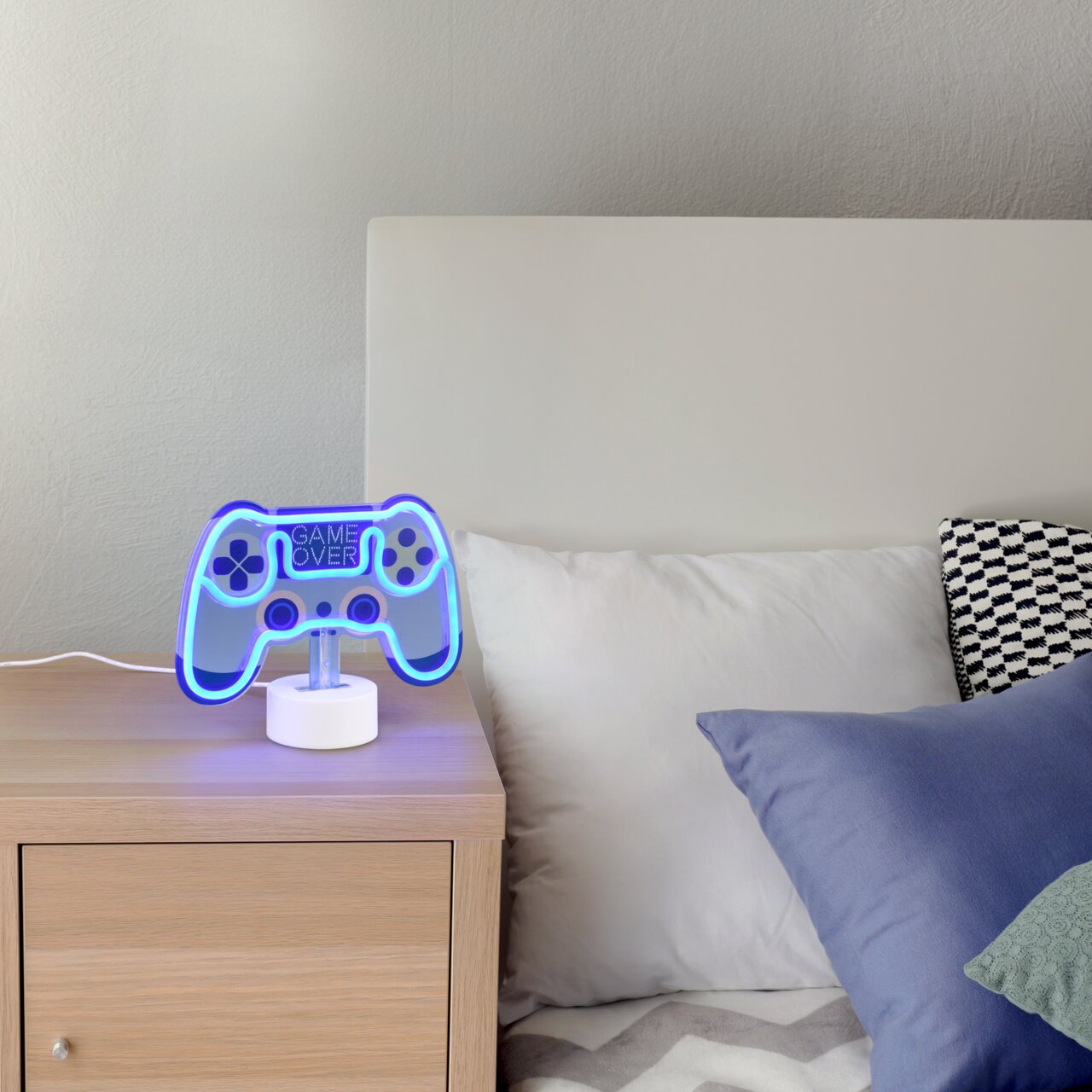 Tischleuchte in Form eines blauen Game-Controllers mit der Aufschrift 'Game Over', auf einem Nachttisch neben einem Bett platziert, seitliche Perspektive.