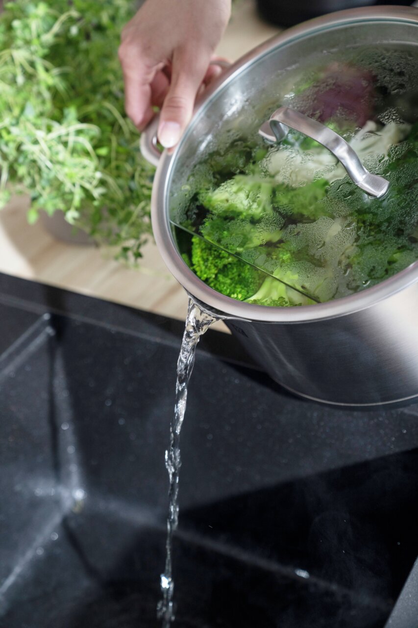 Topf mit Deckel, aus dem Wasser in ein Spülbecken gegossen wird, mit frischem Gemüse im Topf, seitliche Perspektive.