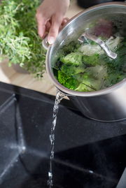 Topf mit Deckel, aus dem Wasser in ein Spülbecken gegossen wird, mit frischem Gemüse im Topf, seitliche Perspektive.
