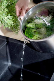 Topf mit Deckel, aus dem Wasser in ein Spülbecken gegossen wird, mit frischem Gemüse im Topf, seitliche Perspektive.