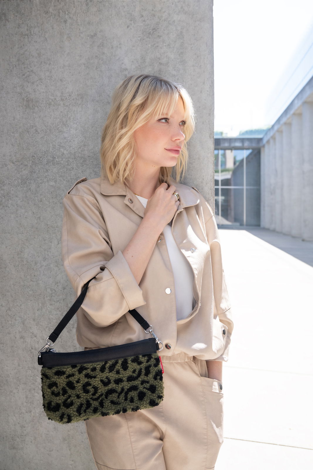 Reisenthel Mini Pouch Teddy Leo Olive Frau in beigem Outfit lehnt an einer Betonwand und hält eine kleine Tasche mit Leopardenmuster in der Hand. Die Tasche hat einen schwarzen Riemen. Seitliche Perspektive.