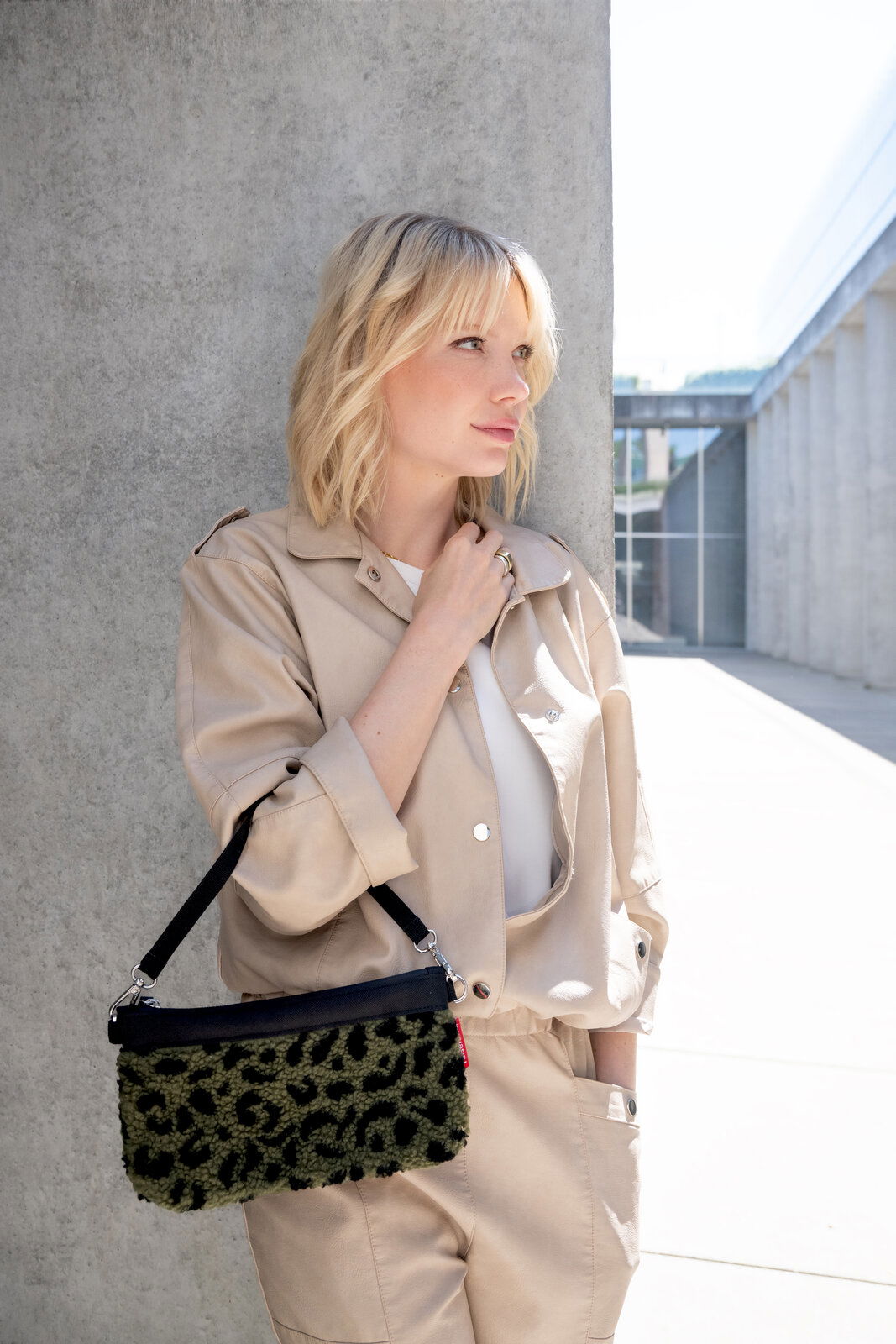 Frau in beigem Outfit lehnt an einer Betonwand und hält eine kleine Tasche mit Leopardenmuster in der Hand. Die Tasche hat einen schwarzen Riemen. Seitliche Perspektive.