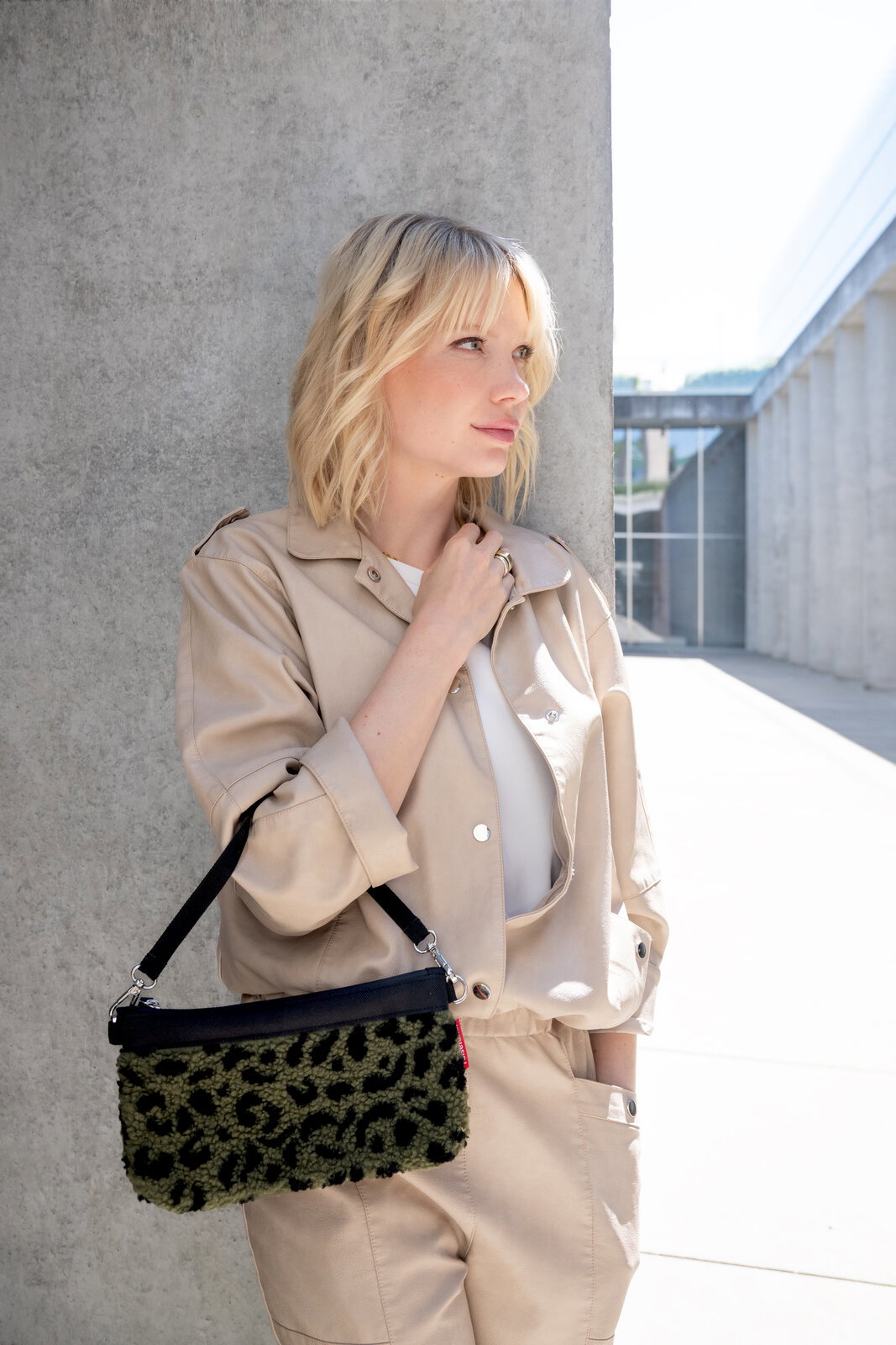 Reisenthel Mini Pouch Teddy Leo Olive Frau in beigem Outfit lehnt an einer Betonwand und hält eine kleine Tasche mit Leopardenmuster in der Hand. Die Tasche hat einen schwarzen Riemen. Seitliche Perspektive.