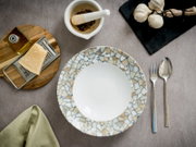 Runder Essteller mit weißer Mitte und Mosaikrand in Beige- und Blautönen auf grauer Tischdecke, umgeben von Besteck, Knoblauch, Mörser und Parmesan – Draufsicht