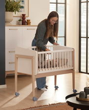 Frau steht neben einem Anstellbett mit einem schlafenden Baby, seitliche Perspektive