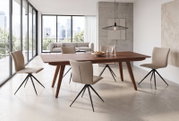 Moderne houten eettafel met donkerbruin tafelblad en schuine poten, omringd door beige gestoffeerde stoelen met zwarte metalen onderstellen, gefotografeerd in driekwart zijaanzicht in een lichte woonkamer met grote ramen.