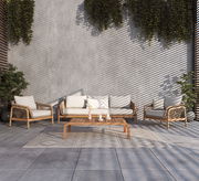 Loungegarnitur-Set aus Holz und Rattan mit beigen Kissen, bestehend aus einem Sofa, zwei Sesseln und einem niedrigen Tisch, auf einer Terrasse mit Pflanzen im Hintergrund, aus frontaler Perspektive.