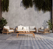 Loungegarnitur-Set aus Holz und Rattan mit beigen Kissen, bestehend aus einem Sofa, zwei Sesseln und einem niedrigen Tisch, auf einer Terrasse mit Pflanzen im Hintergrund, aus frontaler Perspektive.